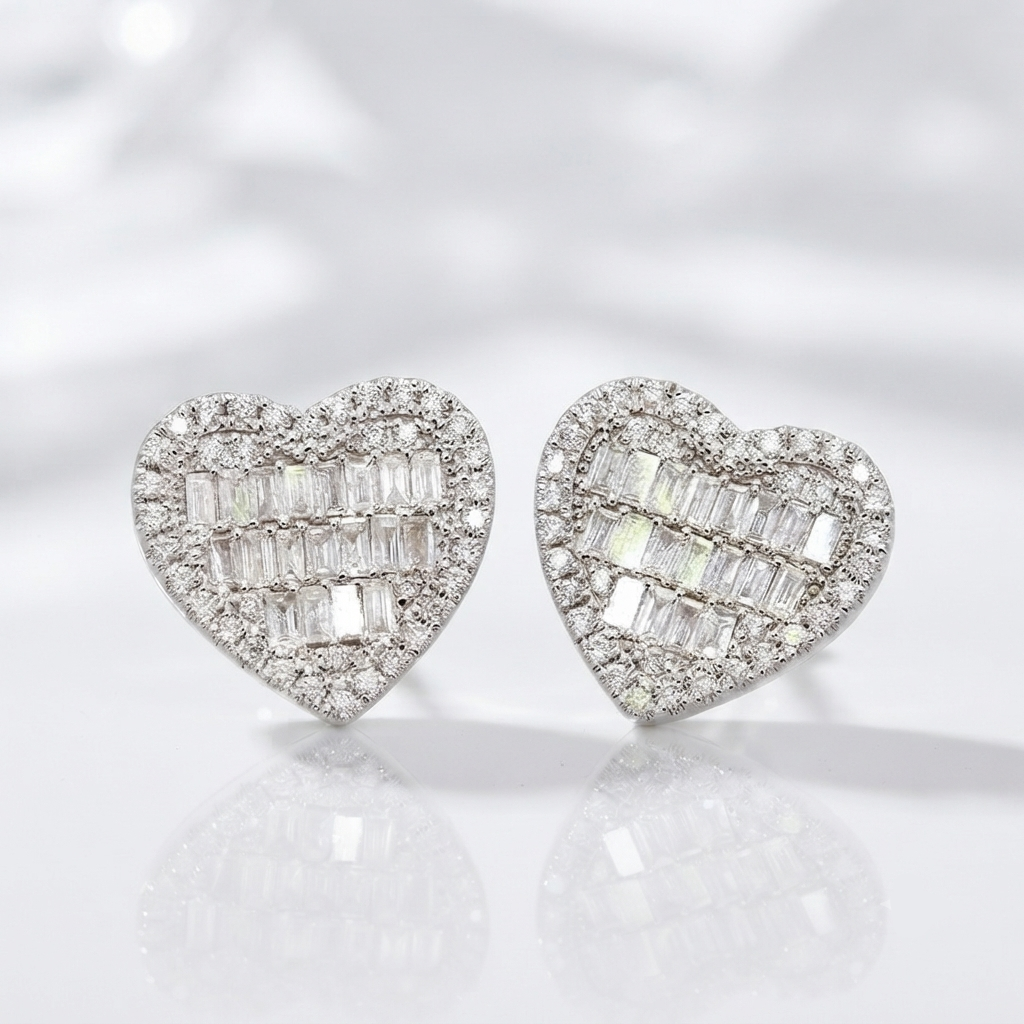 A pair of heart-shaped stud earrings set with baguette-cut clear stones in a silver-toned metal, displayed on a reflective white surface against a soft white background.