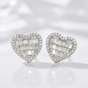 A pair of heart-shaped stud earrings set with baguette-cut clear stones in a silver-toned metal, displayed on a reflective white surface against a soft white background.