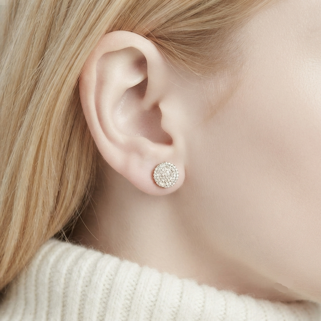 A close-up of a person's ear wearing a small round stud earring with a halo of clear stones in a silver-toned setting, against soft neutral skin tones and blonde hair.