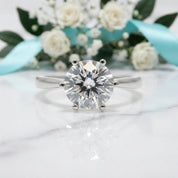 A round brilliant-cut diamond solitaire ring in a silver-colored setting, displayed on a white marble surface with a blurred bouquet of white roses and a light blue ribbon in the background.