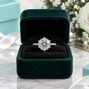 A round-cut diamond solitaire ring in a silver-toned band, displayed inside an open dark green velvet ring box on a marble surface with blurred flowers and a turquoise box in the background.