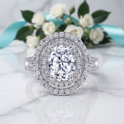 An oval halo ring with a brilliant center stone and surrounding pavé setting, displayed on a white marble surface with white roses and a turquoise ribbon in the background.