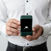 A silver engagement ring with a round diamond set in a dark green presentation box, held by a person wearing a white dress shirt.